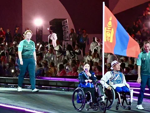 Цэнгэлдэх хүрээлэнгийн тайзан дээр Монгол Улсын төрийн далбааг барьсан хүний ард тэргэнцэртэй хоёр тамирчин, нэг нь цагаан дээл, малгайтай, явж буй нь; цаана нь олон үзэгч