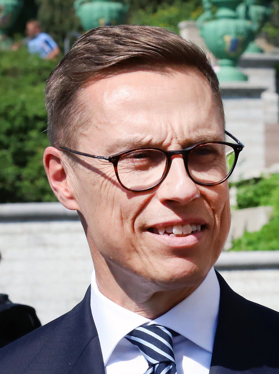 Нүдний шил зүүсэн, хар хөх костюм, судалтай зангиатай Финландын Ерөнхийлөгч Александер Стубб гадаа инээмсэглэн зогсож буй нь