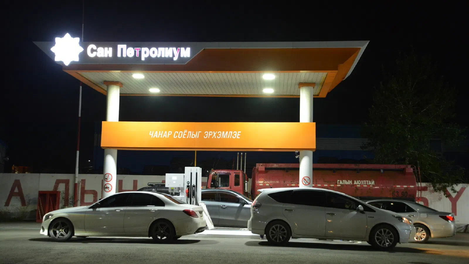 Шөнийн цагаар гэрэлтсэн «Сан Петролиум» бичигтэй ШТС-ын саравчны доор зогсож буй суудлын машинууд, ар талд улаан өнгийн шатахууны цистерн машин
