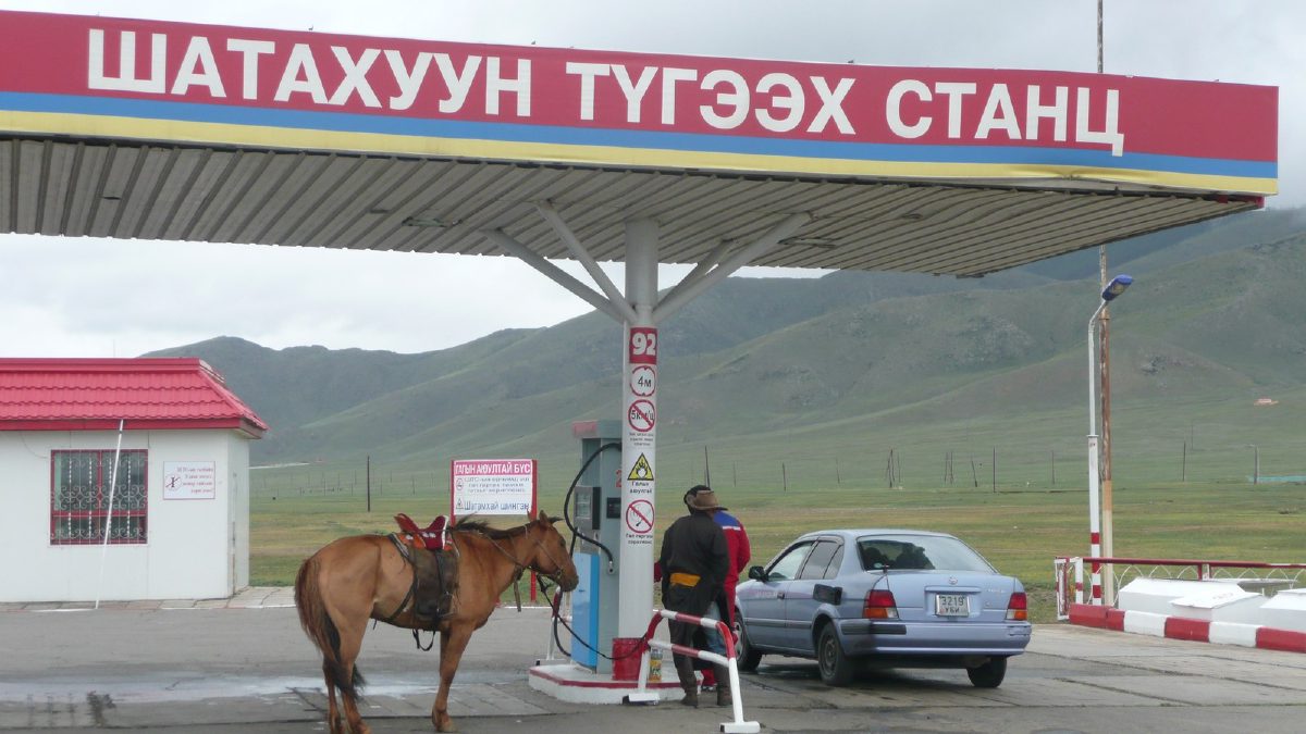 «Шатахуун түгээх станц» гэсэн бичигтэй улаан саравчны доор 92 гэсэн тэмдэгтэй түгээгүүр, эмээллэсэн морь, саарал суудлын машин, ар талд ногоон уул